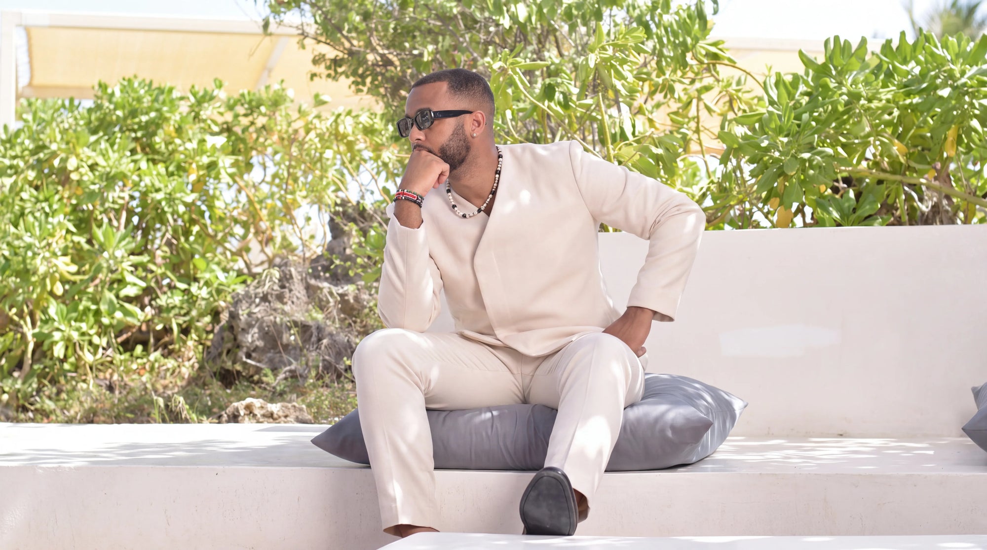 A man wearing a light beige suit and sunglasses sits on a white outdoor bench with gray cushions, surrounded by green foliage.