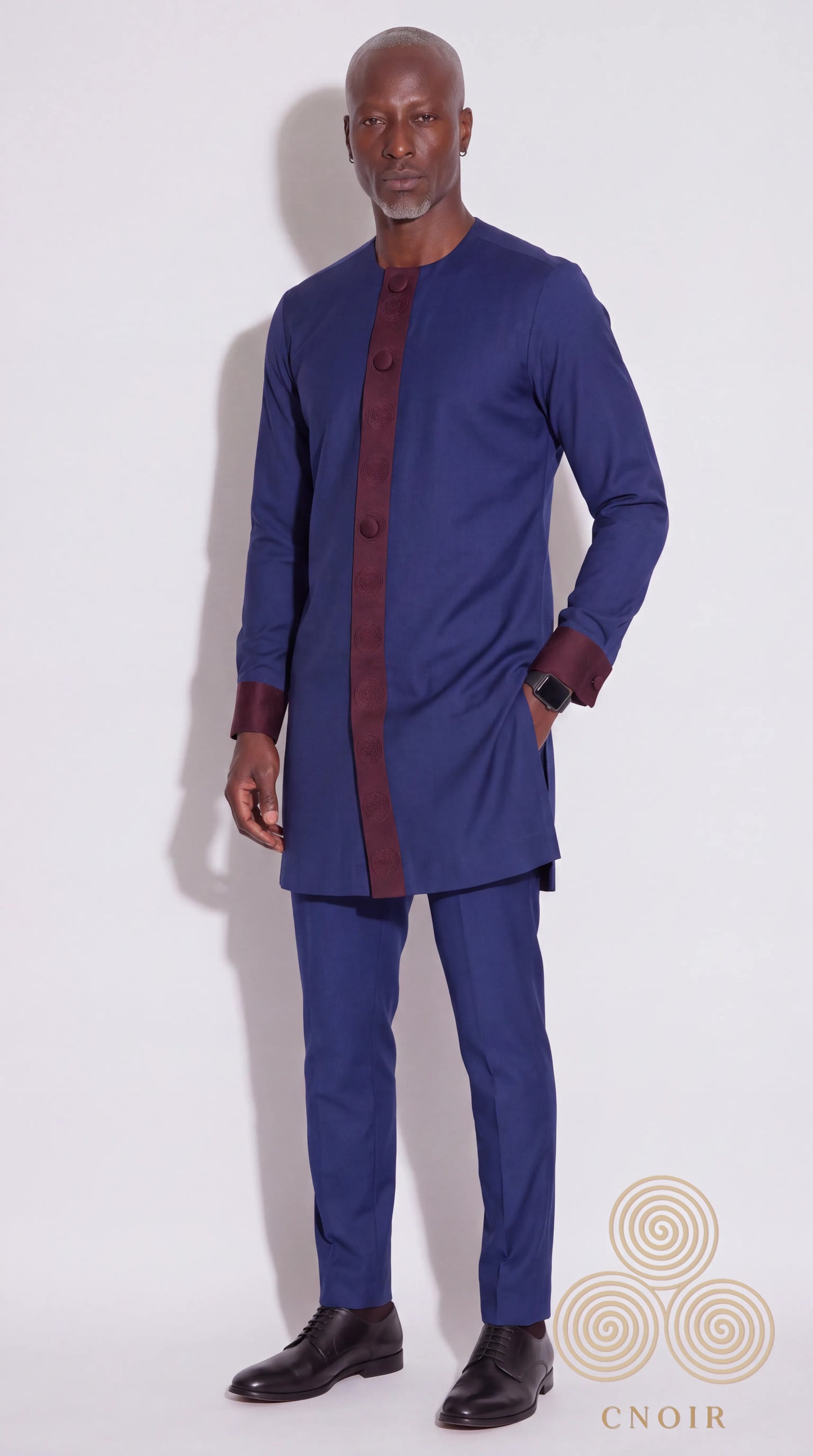 A man modeling a navy blue traditional outfit consisting of a long-sleeve kurta with a contrasting dark red front placket and cuffs, paired with matching navy trousers and black dress shoes, against a plain white studio background.
